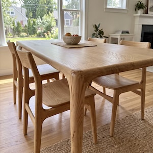 Può includere: Un tavolo da pranzo in legno chiaro con angoli arrotondati e quattro sedie in legno abbinate. Il tavolo presenta una finitura con venature del legno naturali ed è sormontato da una piccola ciotola testurizzata con mele su una tovaglietta.