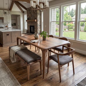 Puede incluir: Una mesa de comedor de madera con bordes redondeados, rodeada de sillas a juego y un banco con un cojín gris. La habitación cuenta con una chimenea de piedra, grandes ventanales y suelos de madera, creando un espacio de comedor acogedor.