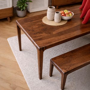 Puede incluir: Una mesa de comedor de madera marrón oscuro con un banco a juego. La mesa tiene una parte superior rectangular y cuatro patas en ángulo. Debajo de la mesa hay una alfombra pequeña. Dos jarrones decorativos y un cuenco de manzanas están sobre la mesa.