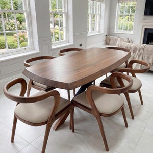 May include: A large, oval-shaped wooden dining table with a natural live edge, supported by a dark metal base. Six chairs with curved wooden frames and light beige upholstered seats are arranged around the table on a white tiled floor.