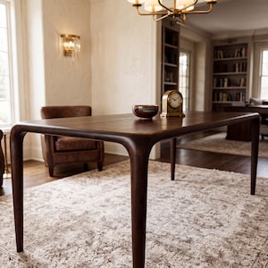 Op de afbeelding: Een donkerbruine houten eettafel met afgeronde hoeken en taps toelopende poten. Een kleine, ronde, donkergekleurde kom en een goudkleurige analoge tafelklok met een witte wijzerplaat en zwarte cijfers staan op het gladde oppervlak van de tafel.