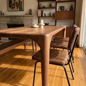 May include: A wooden dining table with rounded corners and a matching bench. Three chairs with brown leather seats and black metal frames are positioned around the table. The room has hardwood floors and a fireplace.
