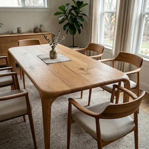 May include: A light oak dining table with six matching chairs. The chairs have light fabric seats and curved wooden backs. A small vase with greenery sits on the table. The table is approximately 200 cm long. The room has a neutral colour palette.