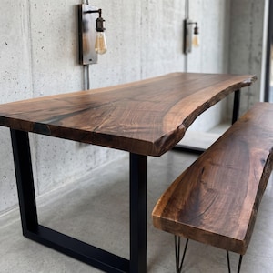 Puede incluir: Un juego de mesa y banco de comedor de madera con diseño de borde vivo. La mesa tiene un acabado marrón oscuro con patas de metal negro. El banco tiene un acabado similar y está soportado por patas de horquilla. El conjunto se exhibe contra una pared de hormigón.