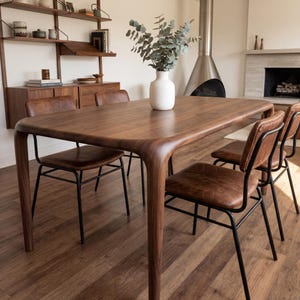 Puede incluir: Una mesa de comedor de madera con bordes redondeados y cuatro sillas. Las sillas tienen asientos de cuero marrón y marcos de metal negro. Un jarrón blanco con vegetación está sobre la mesa. La habitación tiene una estética moderna de mediados de siglo.