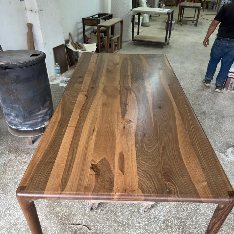 May include: A rectangular wooden dining table with a smooth, polished surface featuring prominent dark brown grain patterns and natural wood knots. The table has rounded corners and four sturdy legs, showcasing a rich, warm wood finish.