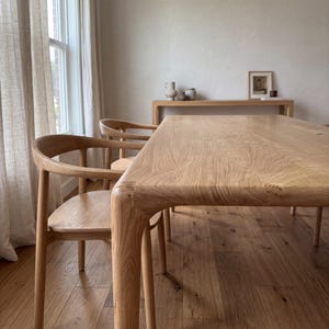 Puede incluir: Una mesa de comedor de madera clara con una superficie rectangular lisa y bordes redondeados, acompañada de sillas de madera a juego con respaldos y reposabrazos curvos, todo con un acabado de veta de madera natural.
