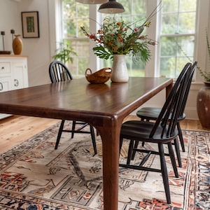 May include: A rectangular dark wood dining table with rounded corners and four tapered legs. Two black spindle-back chairs are positioned at the table. A white ceramic vase with green foliage and red berries sits on the table next to a small wooden bowl.