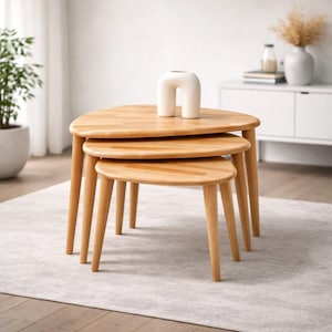 Può includere: Un set di tre tavolini ad incastro con finitura in legno chiaro. Ogni tavolo ha un piano triangolare e gambe affusolate. Un vaso in ceramica bianco a forma di arco è posto sul tavolo più grande. I tavoli sono disposti su un tappeto grigio chiaro.