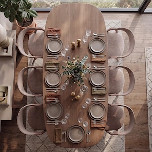Puede incluir: Una mesa de comedor ovalada de madera preparada para siete personas con sillas tapizadas en marrón claro. La mesa incluye platos de cerámica moteada, cubiertos de acero inoxidable, cristalería transparente, servilletas y un centro floral.
