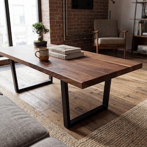 Puede incluir: Una mesa de centro rectangular con un tablero de madera maciza de color marrón oscuro con veta visible y dos patas de metal negro en forma de U. Una taza de cerámica y dos libros apilados se encuentran sobre la superficie de la mesa.