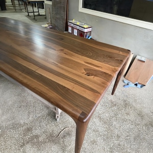 May include: A rectangular dining table crafted from dark brown walnut wood with a smooth, polished surface. The table features a prominent natural wood grain pattern and four tapered legs that extend from the corners of the tabletop.