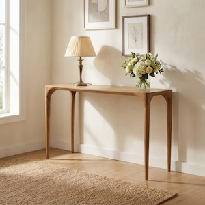 May include: A wooden console table with a curved top and four slender legs. A beige lampshade sits on the table, along with a vase of flowers. The table is placed against a white wall with framed artwork and a window.