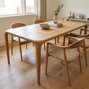 May include: A light-coloured wooden dining table with rounded edges and four matching chairs. The chairs have light-coloured fabric seats and curved wooden backs. A decorative bowl and vase with greenery sit on the table.