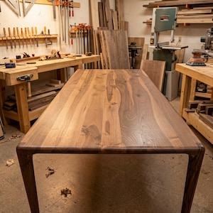 May include: A rectangular dining table crafted from solid walnut wood, featuring a smooth, polished surface with visible natural grain patterns and rounded edges that transition seamlessly into four sturdy, tapered legs.