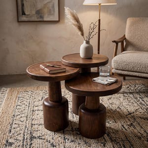 May include: A set of three tiered round side tables crafted from dark brown wood, each featuring a thick cylindrical base and a smooth circular top. The tables are arranged in a cluster, with a small notebook, a white vase, and a glass of water.