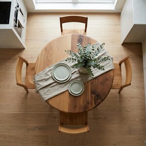 May include: Overhead view of a round wooden dining table with a light-colored runner, two plates, and a vase of greenery. The table is surrounded by four wooden chairs. The floor is made of light-colored wood.
