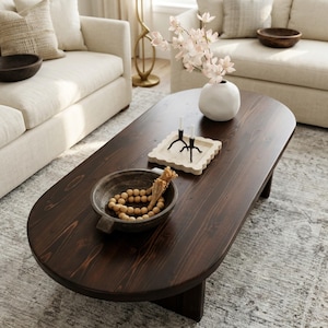 May include: A dark brown oval coffee table with a wooden bowl, decorative tray, and white vase. The table is surrounded by cream-colored sofas and a patterned rug. The vase holds pink flowers, and the tray holds two black candle holders.