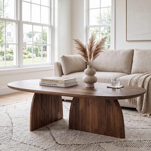May include: An oval-shaped wooden coffee table with a smooth, dark brown finish and two curved, solid panel legs. On the tabletop sit two stacked books titled Mid-Century Modern Design and Architecture of the West, a beige vase, and a glass.