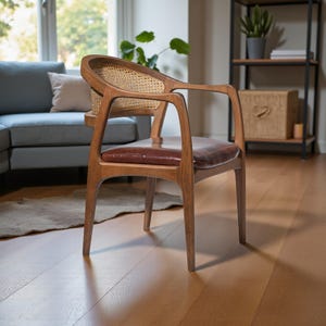 Pode incluir: Poltrona de madeira com encosto em cana tecida e assento em couro castanho. A cadeira tem encosto e apoios de bra&ccedil;os curvos, com acabamento em madeira quente. A cadeira est&aacute; numa sala com ch&atilde;o de madeira e um sof&aacute; cinzento.