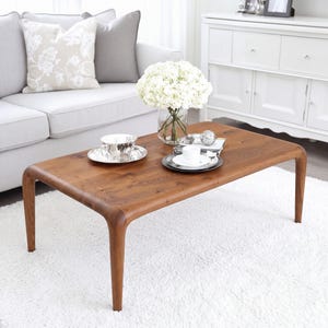 May include: A rectangular wooden coffee table with rounded corners and four tapered legs. The table surface features a natural wood grain finish and holds a glass vase with white flowers, a silver teacup on a saucer, and a white cup on a plate.