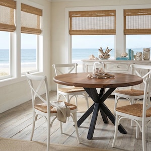 May include: A round wooden dining table with a black metal base, surrounded by white chairs with woven seats. The room has a coastal theme, with natural woven shades and a view of the ocean.