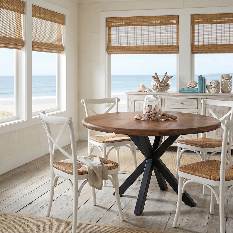 May include: A round wooden dining table with a black metal base, surrounded by white chairs with woven seats. The room has a coastal theme, with natural woven shades and a view of the ocean.