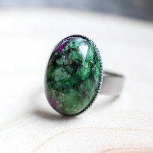 Könnte beinhalten: Ovaler Ring mit einem großen, polierten Edelstein. Der Stein hat eine grüne Basis mit roten Einschlüssen und ist in einem silberfarbenen Metallband mit strukturierter Lünette gefasst. Der Ring wird auf einer hellen Holzoberfläche präsentiert.