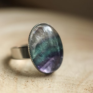 Könnte beinhalten: Ovaler Ring mit einem großen, polierten Fluoritstein in Grün-, Lila- und Weißtönen. Der Stein ist in einem silberfarbenen Metallband mit strukturierter Lünette gefasst. Der Ring wird auf einer hellen Holzoberfläche präsentiert.