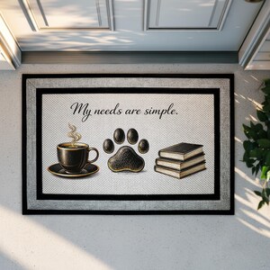 May include: A rectangular doormat with a light-colored textured center and a black border. The text "My needs are simple." is printed above a black coffee cup, a black animal paw print, and a stack of three black books with gold-toned edges.