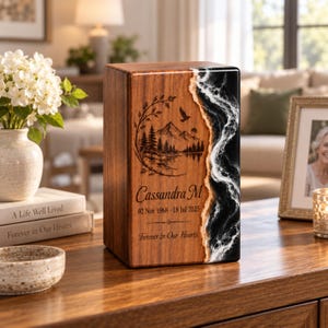 Puede incluir: Una urna funeraria rectangular de madera con un grabado láser de una montaña, un bosque y un pájaro. El frente muestra el nombre Cassandra M, las fechas 02 Nov 1968 - 18 Jul 2025 y el texto Forever in Our Hearts, con un borde de resina blanco y negro.