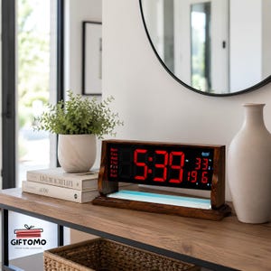 May include: A digital clock with a dark wood frame and a black display screen featuring bright red LED numbers. The display shows the time, the day of the week, the temperature in Celsius, and the date. The clock sits on a flat wooden base.