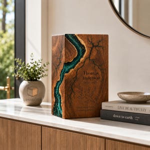 Op de afbeelding: Een rechthoekige houten urn met een centrale inleg van blauwgroene hars met gouden accenten. De voorzijde is gegraveerd met de naam Thomas Anderson, de jaren 1918-2023, een botanische lijntekening en de tekst Always in our heart, forever in our love.