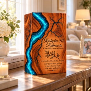 May include: A rectangular wooden memorial urn featuring a vibrant, glowing blue resin river inlay with fractal patterns. The front is engraved with the name Kristopher M. Palmaccio, dates, a leaf branch graphic, and a sentimental poem in black text.