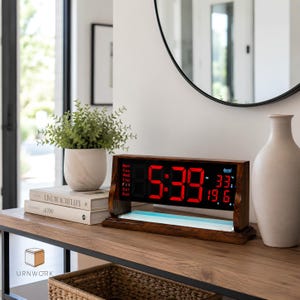 Puede incluir: Un reloj digital rectangular con marco de madera oscura y pantalla negra. La pantalla muestra la hora 5:39 en grandes dígitos LED rojos, junto con el día de la semana, la temperatura en grados Celsius y otros indicadores en texto rojo.