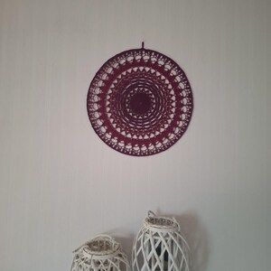 Può includere: Un arazzo da parete circolare bordeaux all'uncinetto con un design traforato dettagliato. Sotto, due lanterne bianche intrecciate con manici in corda poggiano su una superficie bianca. Le lanterne hanno un motivo a griglia.