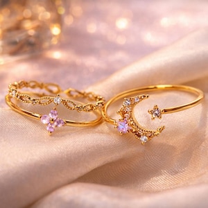 Può includere: Due anelli color oro con disegni celesti poggiano su una superficie satinata. Uno presenta un design a mezzaluna e stella, l'altro un design floreale e diamantato. Gli anelli sono delicati ed eleganti.