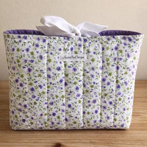 May include: A rectangular fabric tote bag with a floral pattern of purple flowers and green leaves on a white background. The bag has a purple interior and a white bow. The label reads "JaideByDesign".