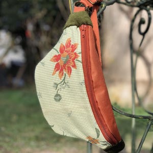 Peut inclure: Sac banane floral avec un motif à carreaux verts et blancs et une grande fleur orange. Le sac a une fermeture éclair et une sangle de couleur rouille, avec une bordure verte. Le sac est suspendu à un support en métal décoratif.