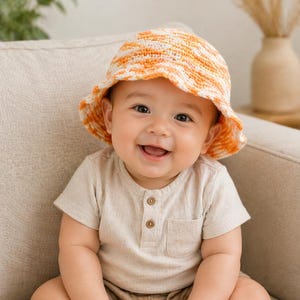 May include: A smiling baby wearing a crocheted orange and white sun hat and a beige short-sleeved shirt with a pocket. The baby is sitting on a beige couch.