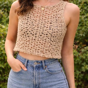 Könnte beinhalten: Beige gehäkeltes Crop-Top mit floralem Muster. Das Top ist ärmellos und hat einen runden Ausschnitt. Das Model trägt hellblaue Jeans. Der Hintergrund ist ein grüner Busch.