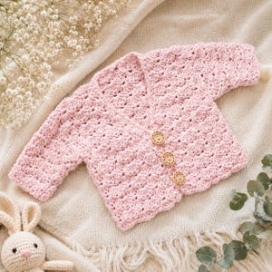 May include: A pale pink, crocheted baby cardigan with three wooden buttons. The handmade sweater has a textured stitch pattern and a scalloped edge. The cardigan is displayed on a cream-coloured blanket, with a stuffed bunny toy in the corner.