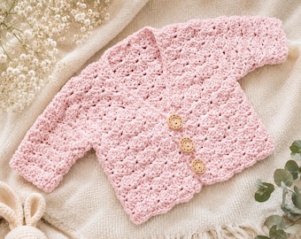 May include: A light pink, crocheted baby cardigan with three wooden buttons. The handmade sweater features a textured stitch pattern and a scalloped edge. The cardigan is displayed on a cream-colored blanket, with a stuffed bunny toy in the corner.