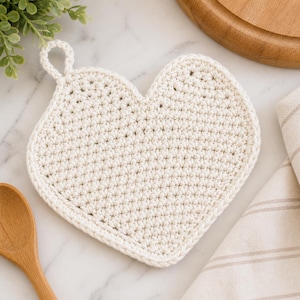 Può includere: Presina a forma di cuore all'uncinetto color crema, con un anello per appenderla. La presina è realizzata con filato a trama fitta e i bordi sono rifiniti con cura. Un cucchiaio di legno e un canovaccio di lino a righe sono sullo sfondo.