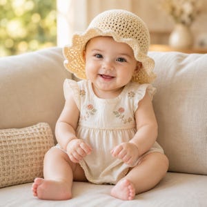 May include: A smiling baby wearing a cream-colored crochet sun hat and a matching romper with floral embroidery. The baby is sitting on a beige couch, with a neutral background.