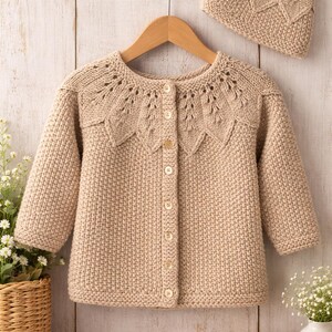 May include: A beige knitted baby cardigan and matching hat set. The cardigan has a round neckline with decorative openwork and is buttoned down the front. The hat has a leaf-like pattern. The set is displayed on a wooden hanger.