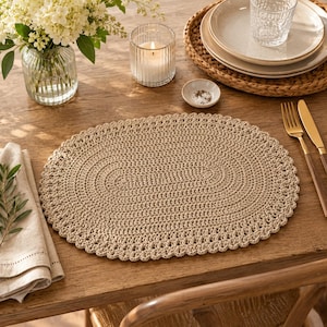 Puede incluir: Un mantel individual ovalado beige tejido a ganchillo sobre una mesa de madera. El mantel individual tiene una superficie texturizada y un borde festoneado. Otros elementos en la mesa incluyen una vela, platos, un vaso, cubiertos y un jarrón con flores blancas.