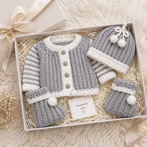 May include: A grey and white crochet baby set, comprising a cardigan, hat, and booties, presented in a gift box. The set features a button-down cardigan, a hat with pom-poms, and matching booties. The set is handmade with love.