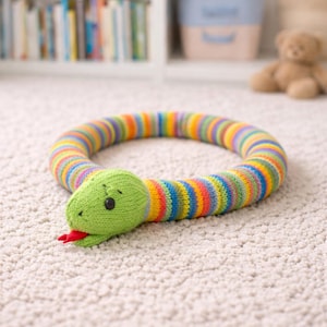 Könnte beinhalten: Eine gestrickte Spielzeugschlange mit einem grünen Kopf und einem bunten gestreiften Körper. Der Schlangenkopf hat schwarze Knopfaugen und eine rote Zunge. Der Körper ist ein Ring aus Regenbogenfarben.