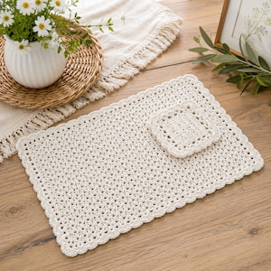 May include: Cream-colored crochet placemat and coaster set. The rectangular placemat and square coaster have a scalloped edge. The open weave design is visible. The set is on a wooden surface, with a vase of flowers and a framed picture in the background.